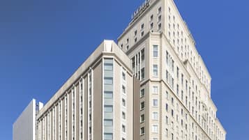 A large, multi-story hotel with a prominent corner entrance, a clear sky, and a few trees in front.