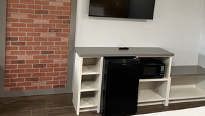 Ein Zimmer mit Backsteinwand, einem an der Wand befestigten Flachbildfernseher, einem weißen Schreibtisch mit schwarzem Kühlschrank und einer Mikrowelle sowie einem Holzboden.