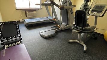A gym with treadmills, an elliptical, and a stationary bike. There is a purple exercise mat and a small table with a laptop.