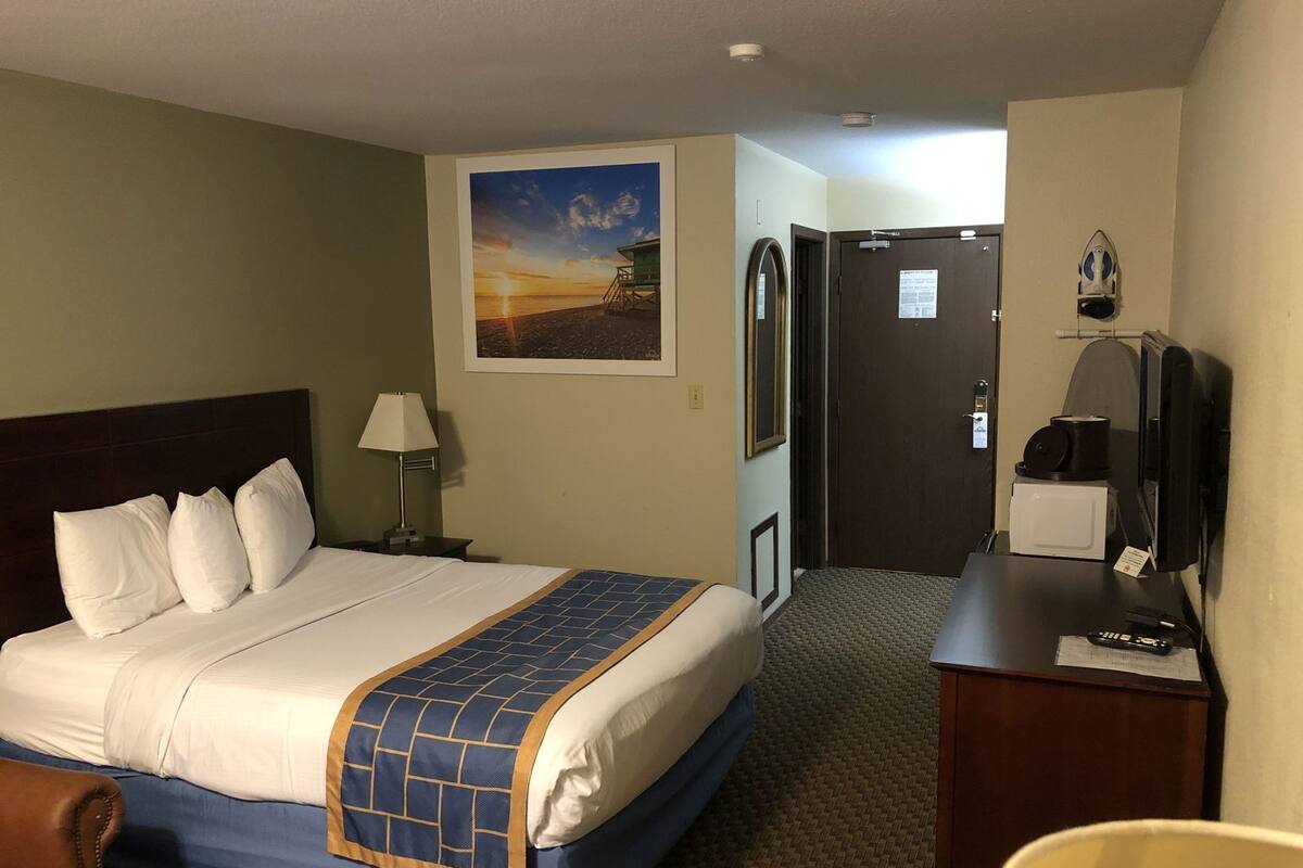 Habitación de hotel con cama, escritorio con computadora, televisor, puerta y un cuadro en la pared.