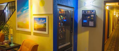 Um corredor de hotel com uma máquina de vendas automática, uma cadeira e quadros nas paredes.