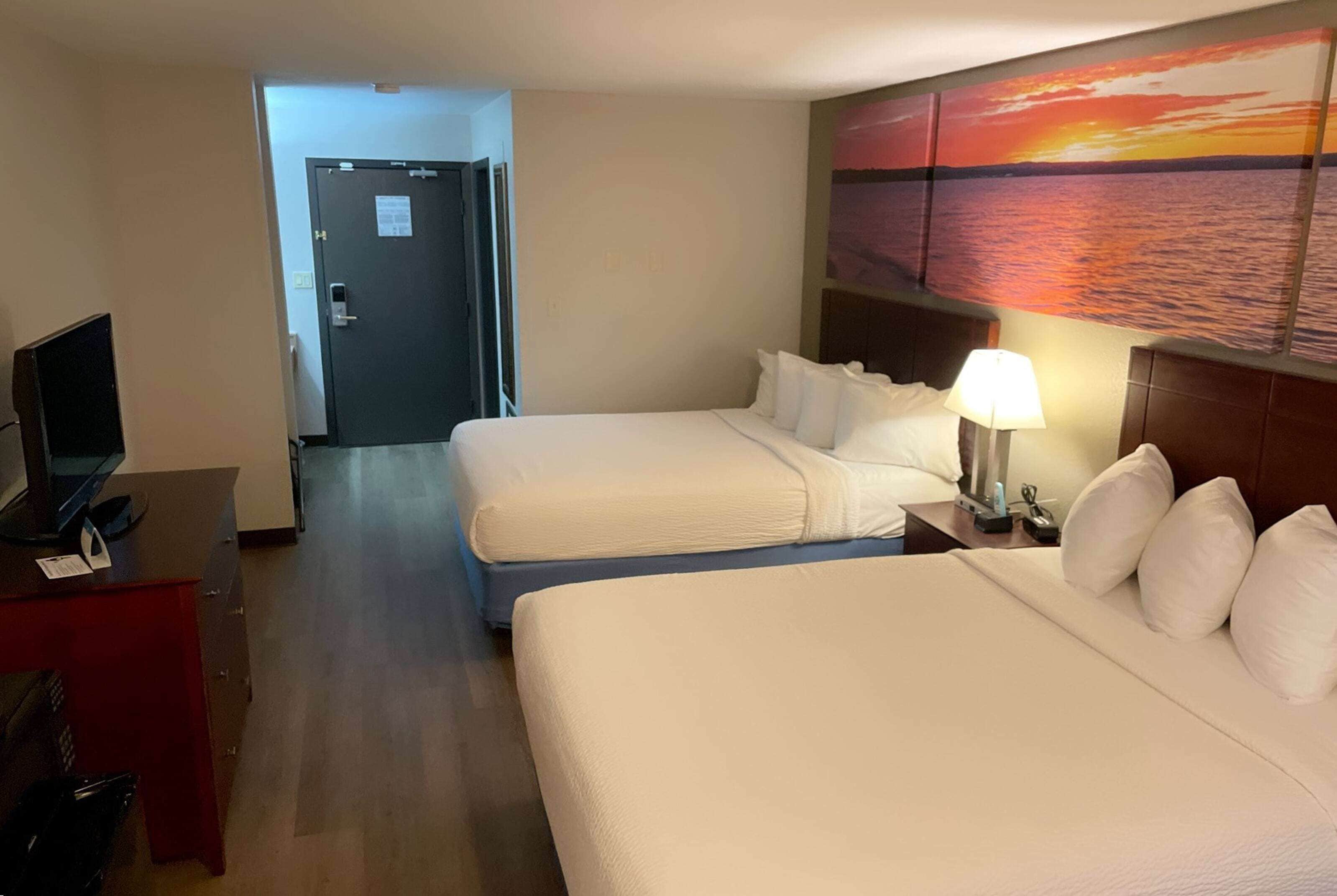 Habitación de hotel con dos camas, un escritorio con computadora, televisor y un cuadro de un atardecer sobre el agua en la pared.