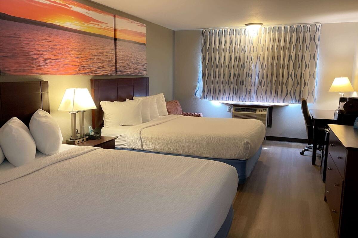 Habitación de hotel con dos camas, un escritorio, una silla, una lámpara y un cuadro grande con un atardecer sobre el agua.