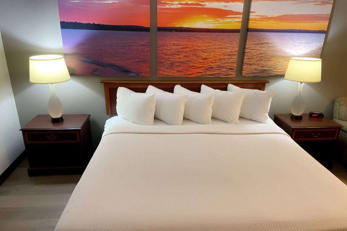 Habitación de hotel con una cama grande, dos mesitas de noche con lámparas, y un cuadro de un atardecer sobre el agua en la pared.