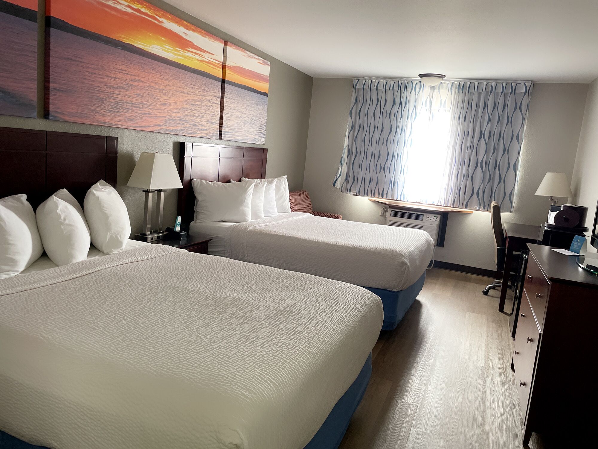 A hotel room with two beds, a desk, a chair, a lamp, a window with curtains, and a painting of a sunset.