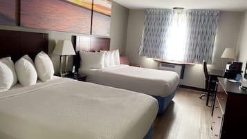 A hotel room with two beds, a desk, a chair, a lamp, a window with curtains, and a painting of a sunset.