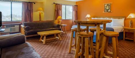 A hotel room with a sofa, a small table, wooden stools, a bed, and a window with curtains.