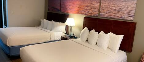 A hotel room with two beds, a nightstand with a lamp, and a large sunset painting on the wall.