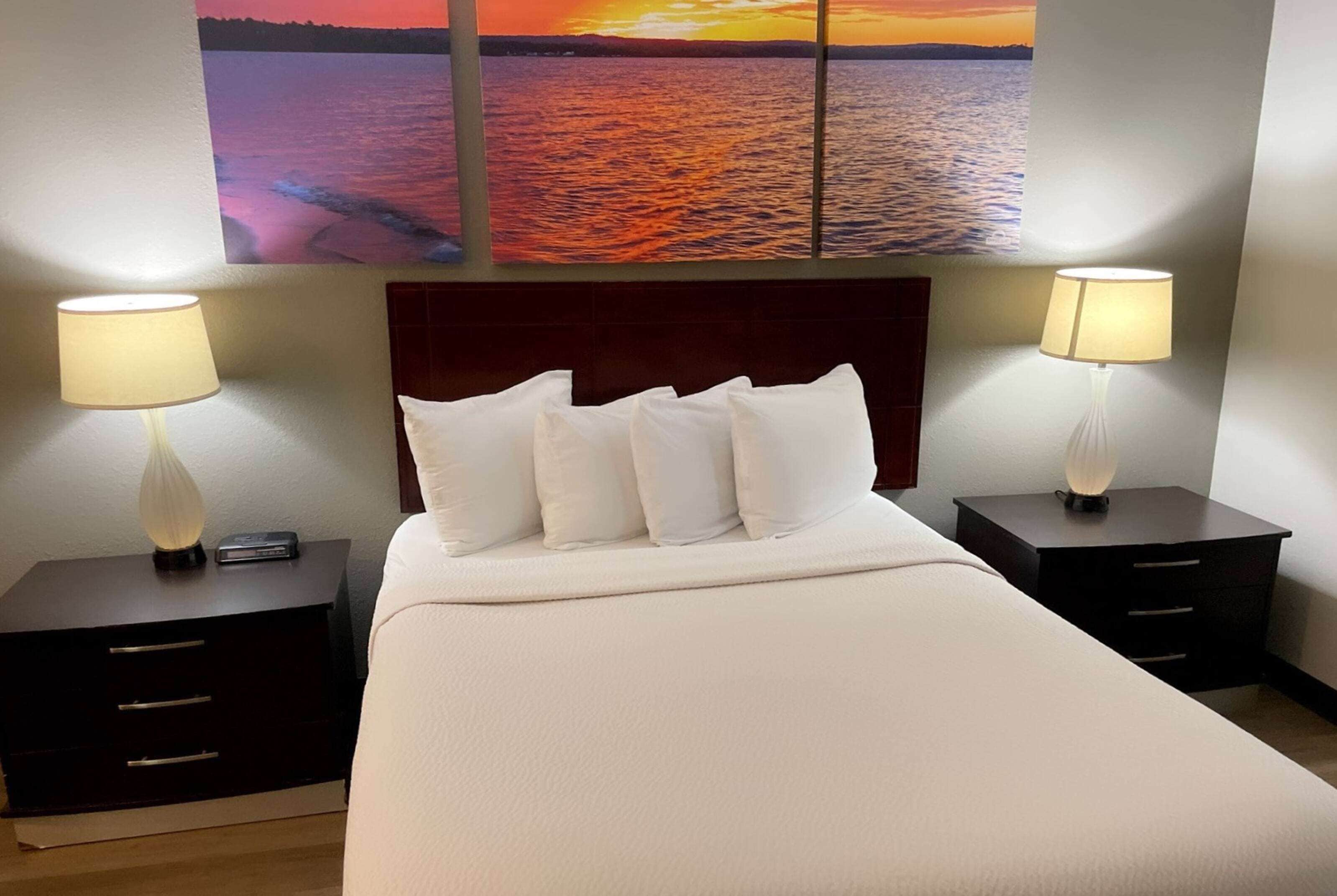 A hotel room with a large bed, two bedside tables with lamps, a headboard, and a painting of a sunset over water on the wall.