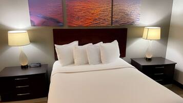 A hotel room with a large bed, two bedside tables with lamps, a headboard, and a painting of a sunset over water on the wall.