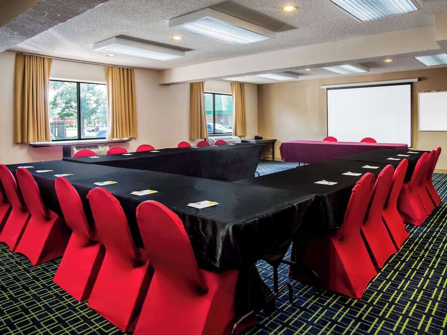Un salón de conferencias con una mesa larga, sillas rojas, una pantalla de proyector y ventanas grandes con cortinas.