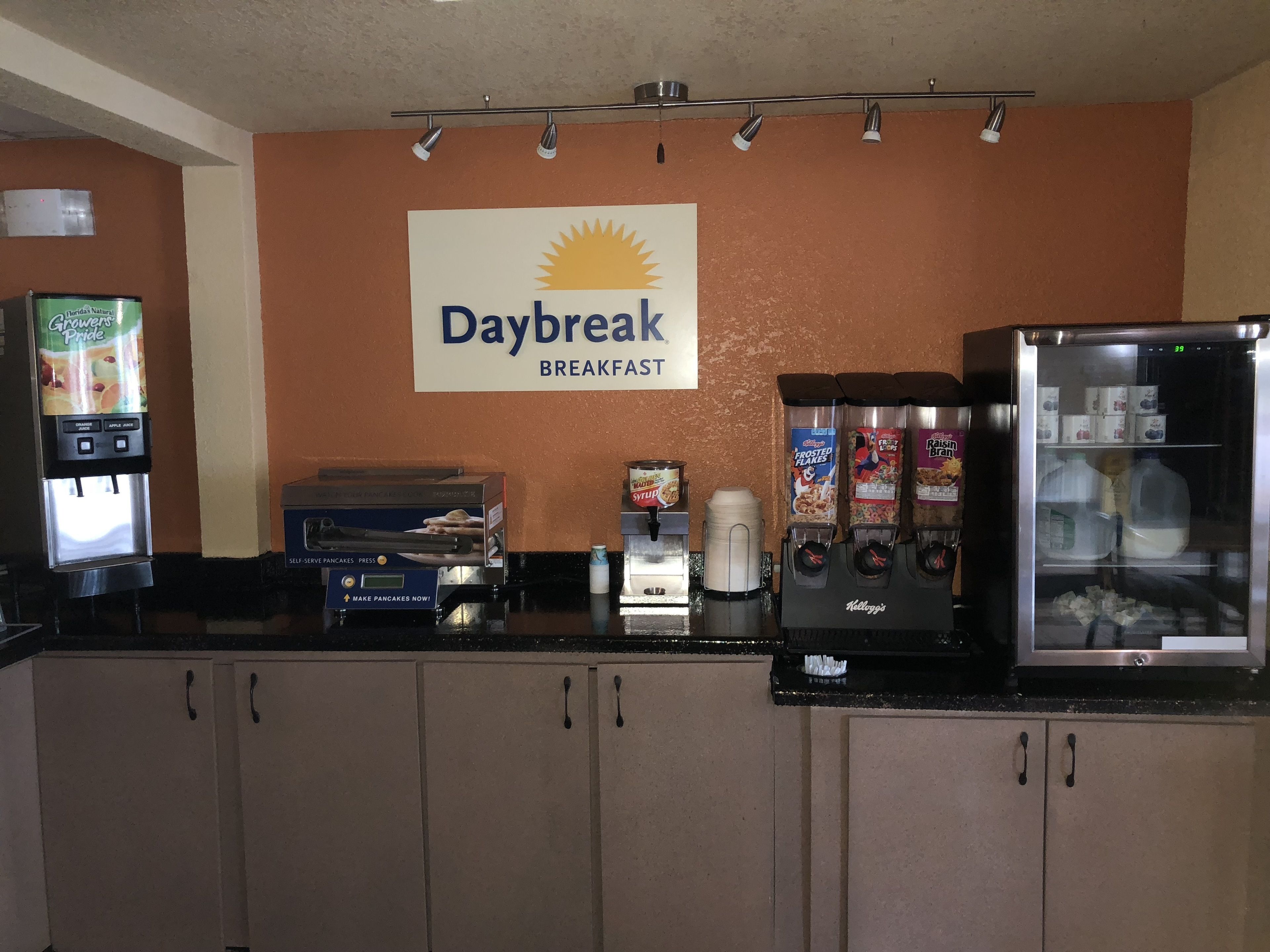 Ein Frühstücksbereich mit einem Daybreak-Schild, Selbstbedienungs-Kaffeemaschinen, einem Getreideautomaten und einem Kühlschrank.