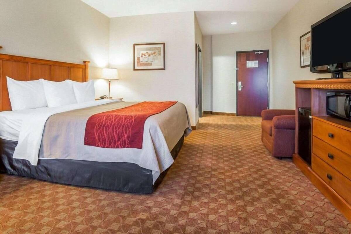 A hotel room with a bed, a desk with a lamp, a television, and a door.