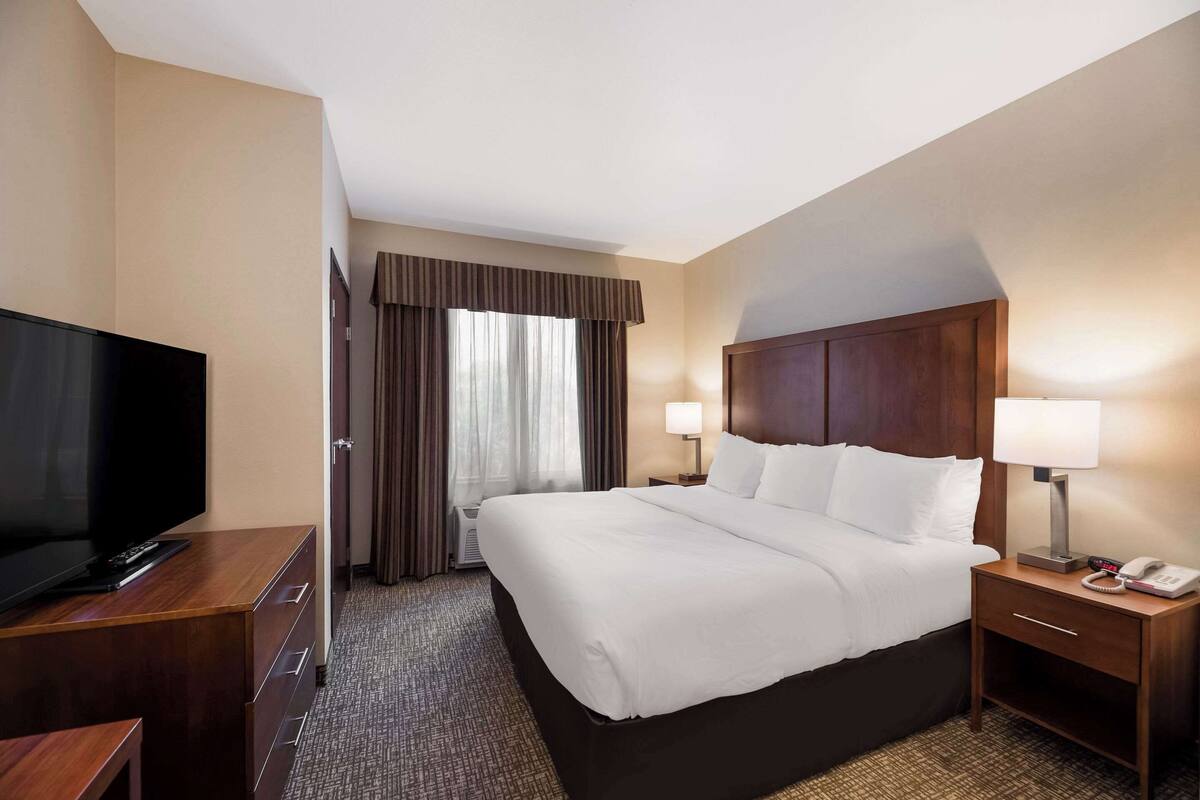 A hotel room with a large bed, a TV on a wooden dresser, a nightstand with a lamp, and a window with curtains.
