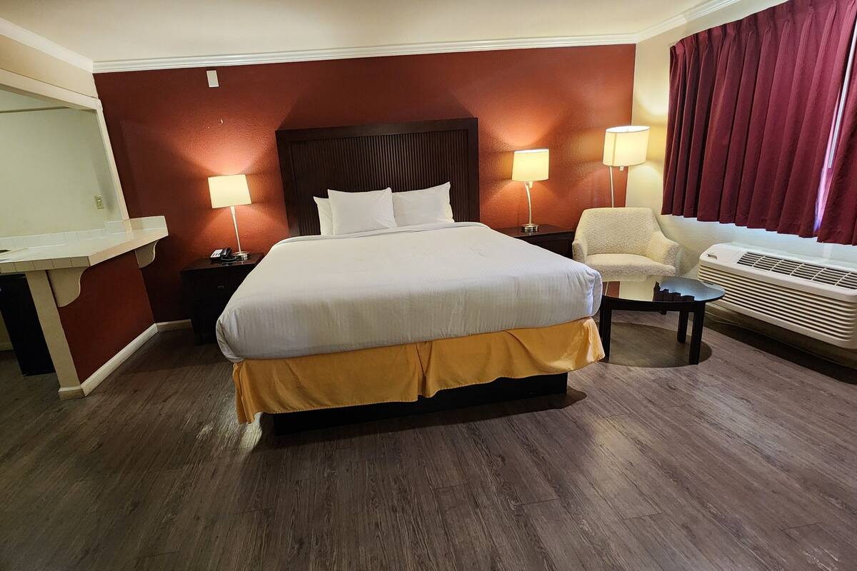 A hotel room with a large bed, bedside lamps, a desk, a chair, and an air conditioning unit.