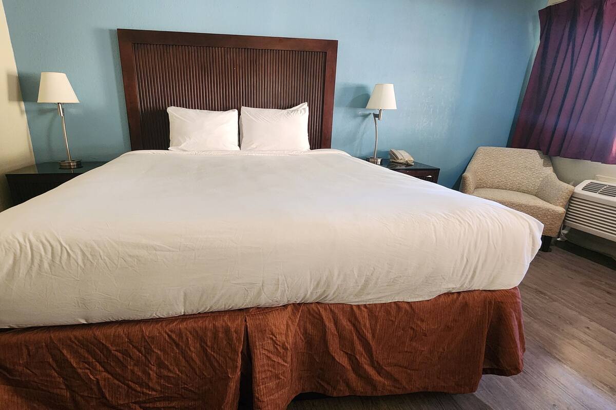 A large bed with a wooden headboard, two bedside lamps, a chair, and an air conditioning unit.