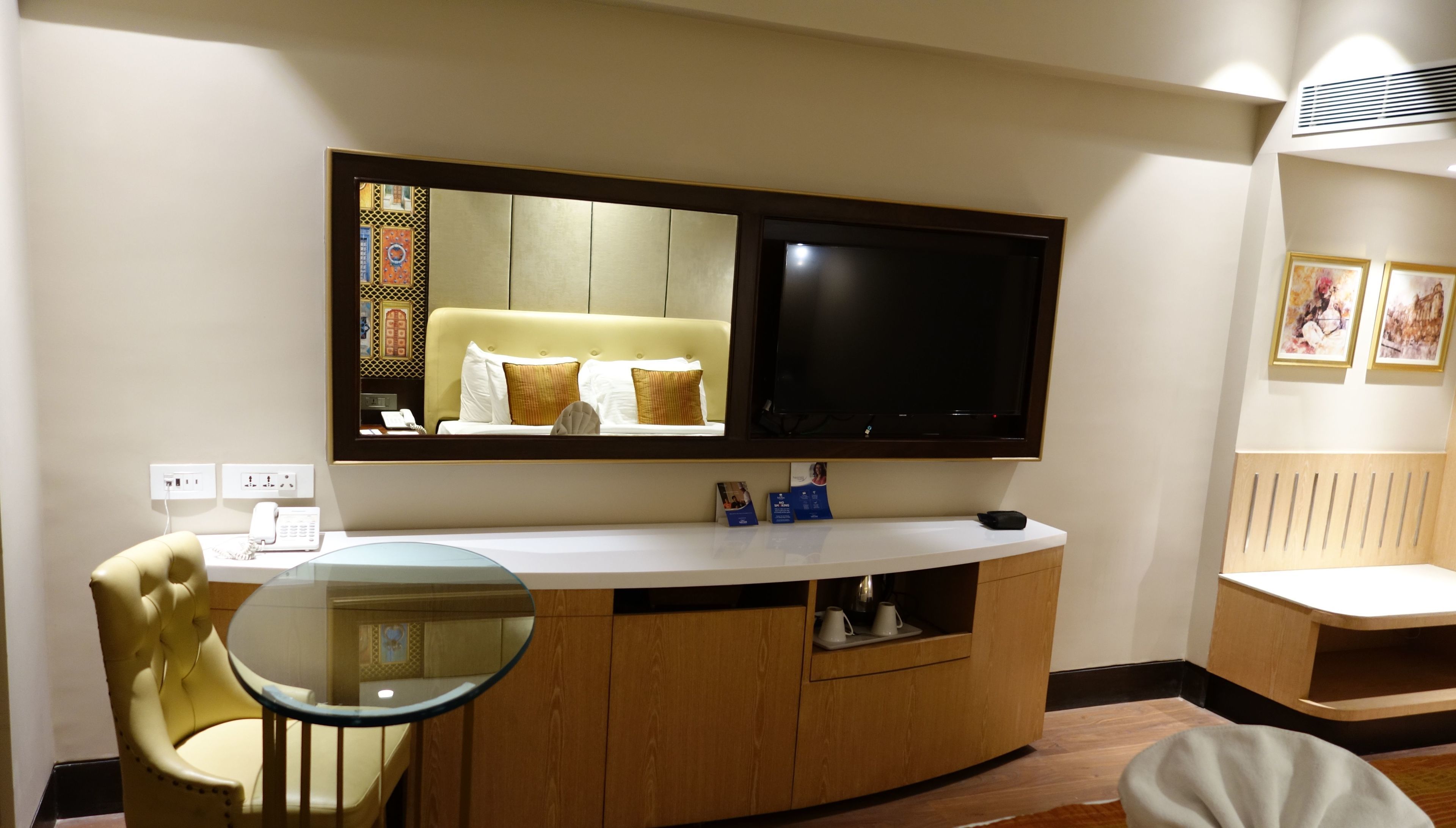 Ein Hotelzimmer mit einem großen Spiegel, einem Flachbildfernseher, einem runden Glastisch, einem gelben Sessel und einem Holzkasten.