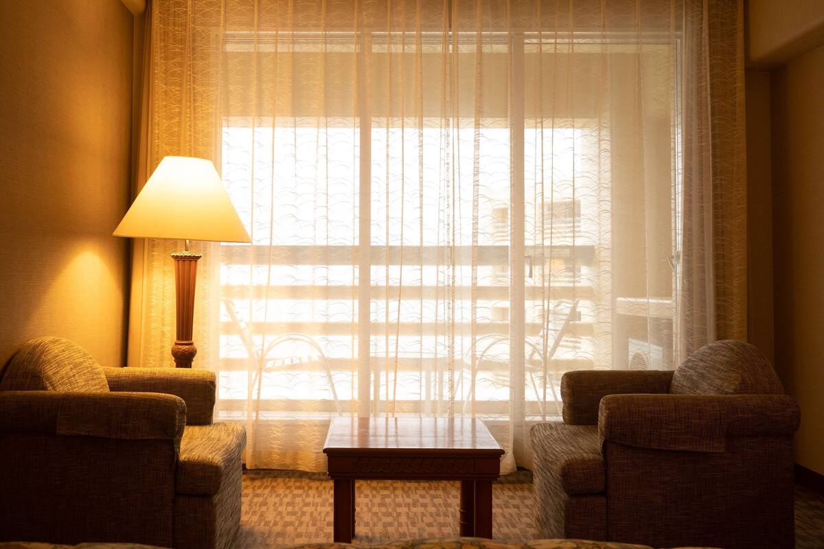A hotel room with a large window, a lamp, two armchairs, and a small table.