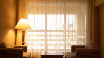A hotel room with a large window, a lamp, two armchairs, and a small table.