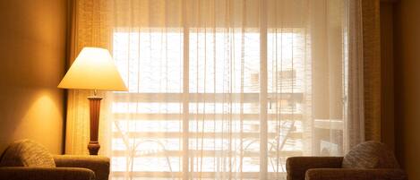 A hotel room with a large window, a lamp, two armchairs, and a small table.