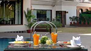 A dining table with breakfast items and drinks, a pool, and a building with a red awning and windows in the background. - Park Plaza Jodhpur (Jodhpur)