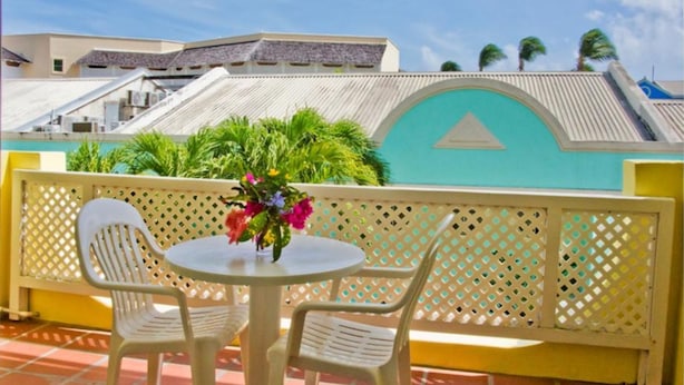 A balcony with two white plastic chairs and a small round table with a vase of flowers. The balcony has a yellow railing and a turquoise wall with a triangular design. - Meridian Inn (St. Lawrence Gap)