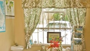 A small room with a window, a sink, a table with a checkered tablecloth, and a bed with a green blanket. - Meridian Inn (St. Lawrence Gap)