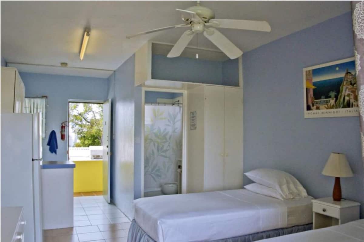 A hotel room with a bed, a nightstand, a ceiling fan, a window with a view, and a door leading to a bathroom.