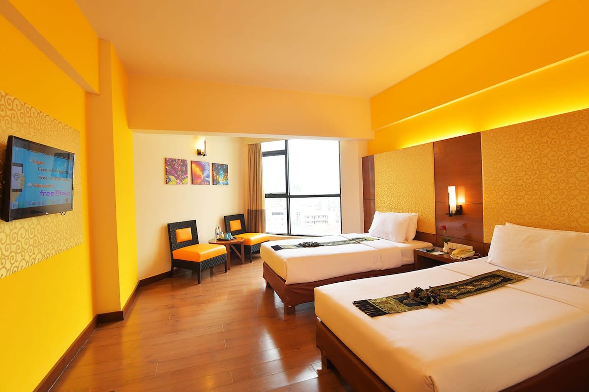 A hotel room with two beds, a desk, chairs, and a TV displaying Wi-Fi information.