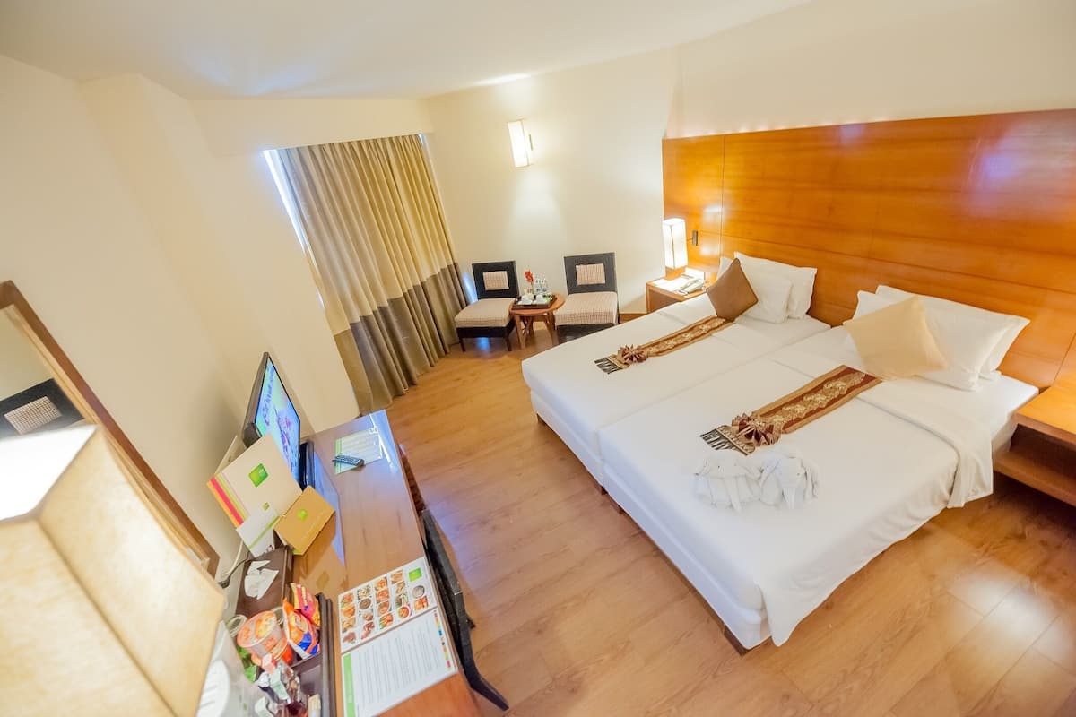 Hotellihuone, jossa on suuri sänky, työpöytä tietokoneella, kaksi tuolia, pieni pöytä lampun kanssa ja ikkuna, jossa on verhot.
