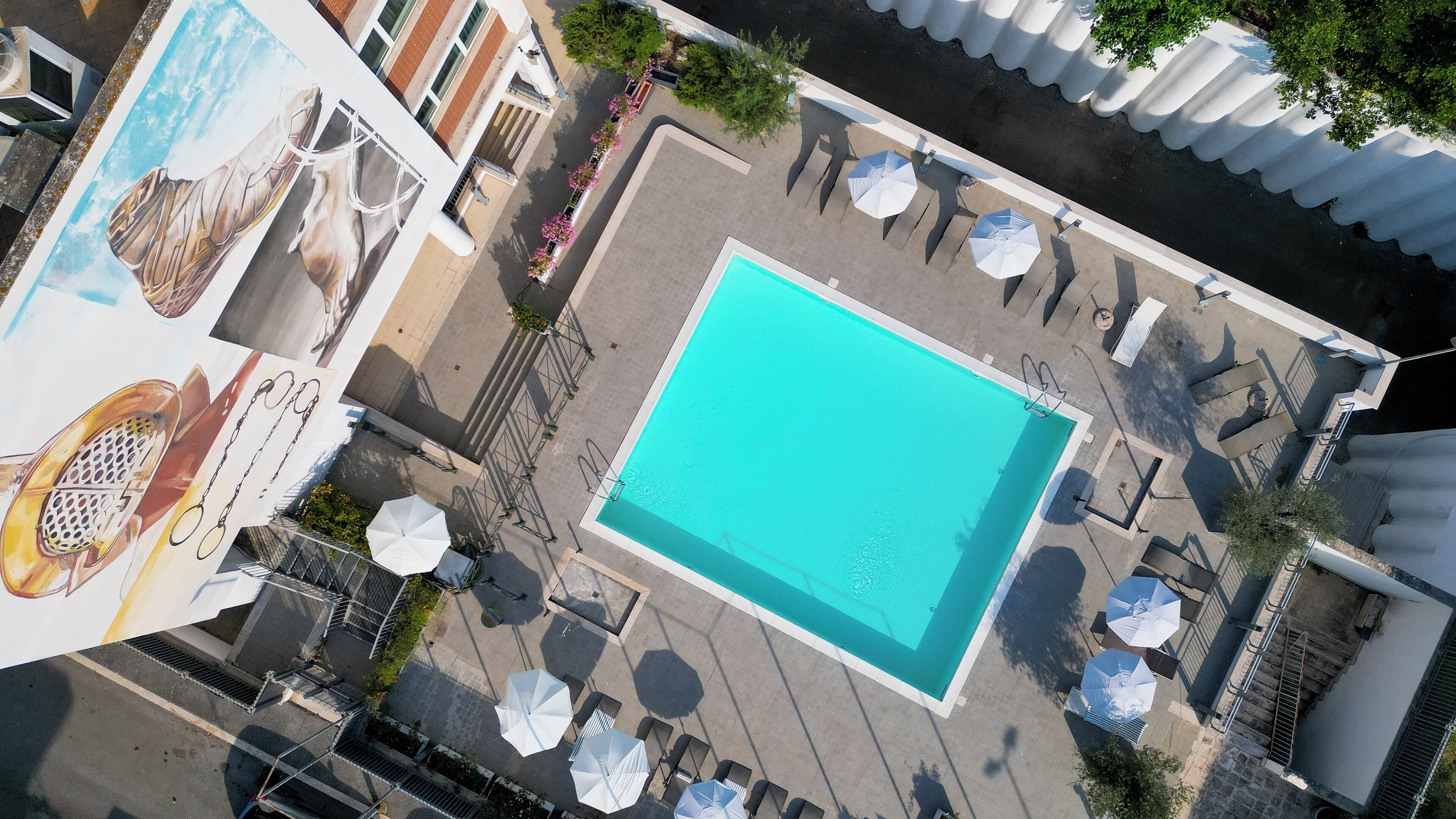 An aerial view of a rooftop pool area with a rectangular pool, surrounded by lounge chairs and umbrellas, and a mural on the left side.
