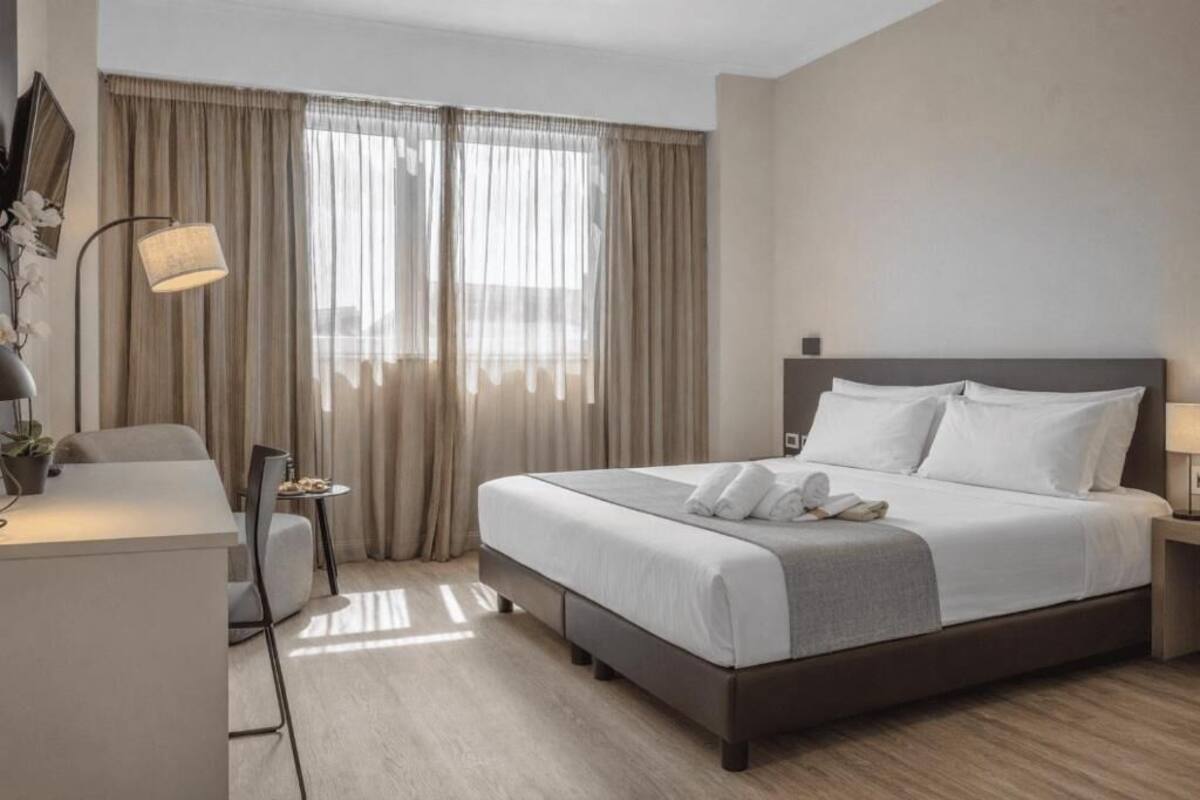 A hotel room with a bed, bedside table, lamp, chair, and a desk with a plant.