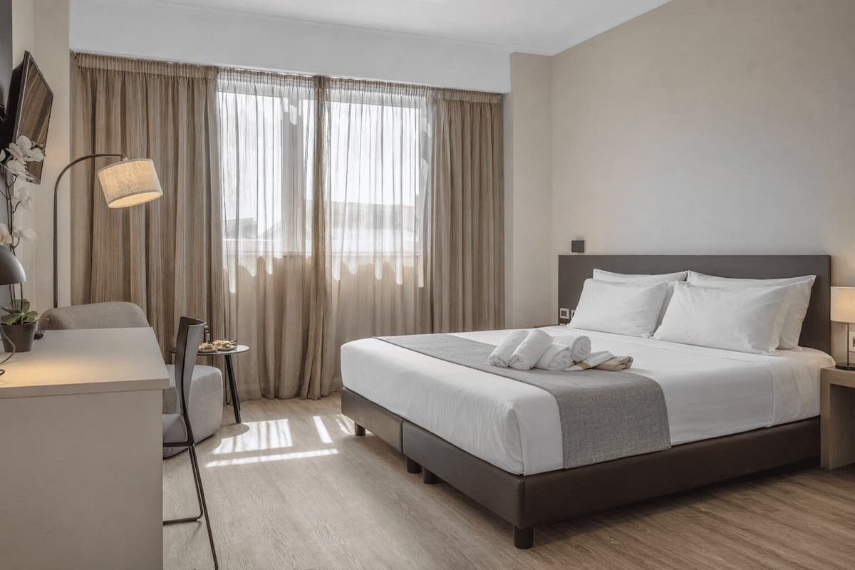 Ein Hotelzimmer mit einem großen Bett, einem Schreibtisch, einem Stuhl, einer Lampe und einem fenster mit Vorhängen.