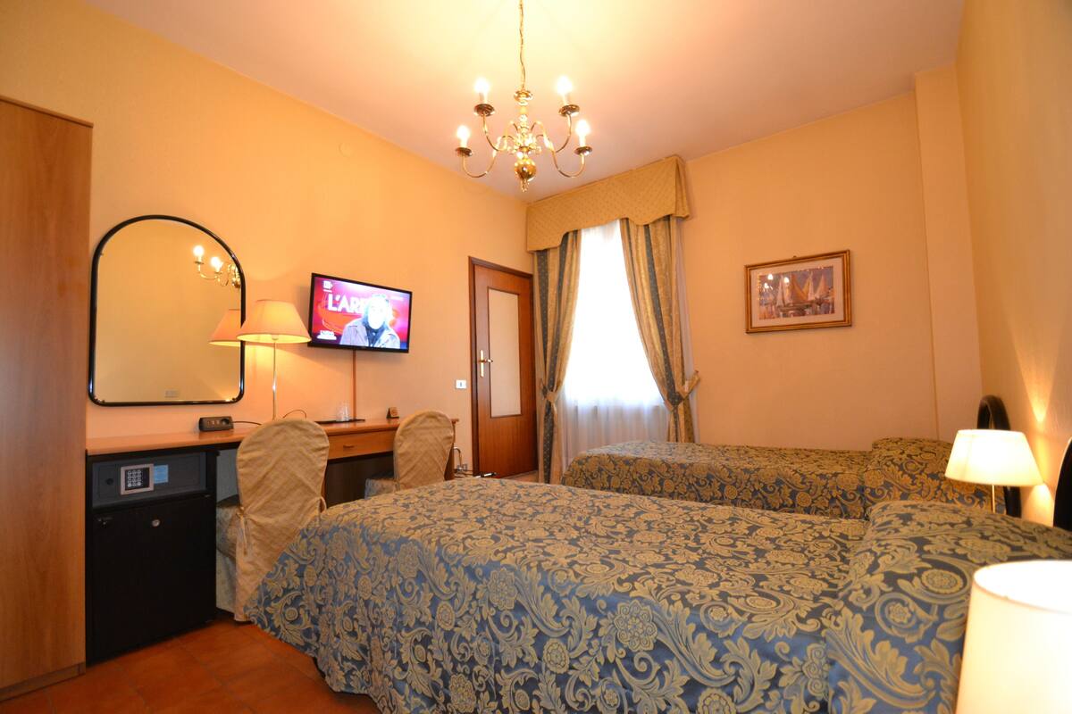 Une chambre d’hôtel avec deux lits, un bureau avec un ordinateur, une télévision, un miroir et un lustre.