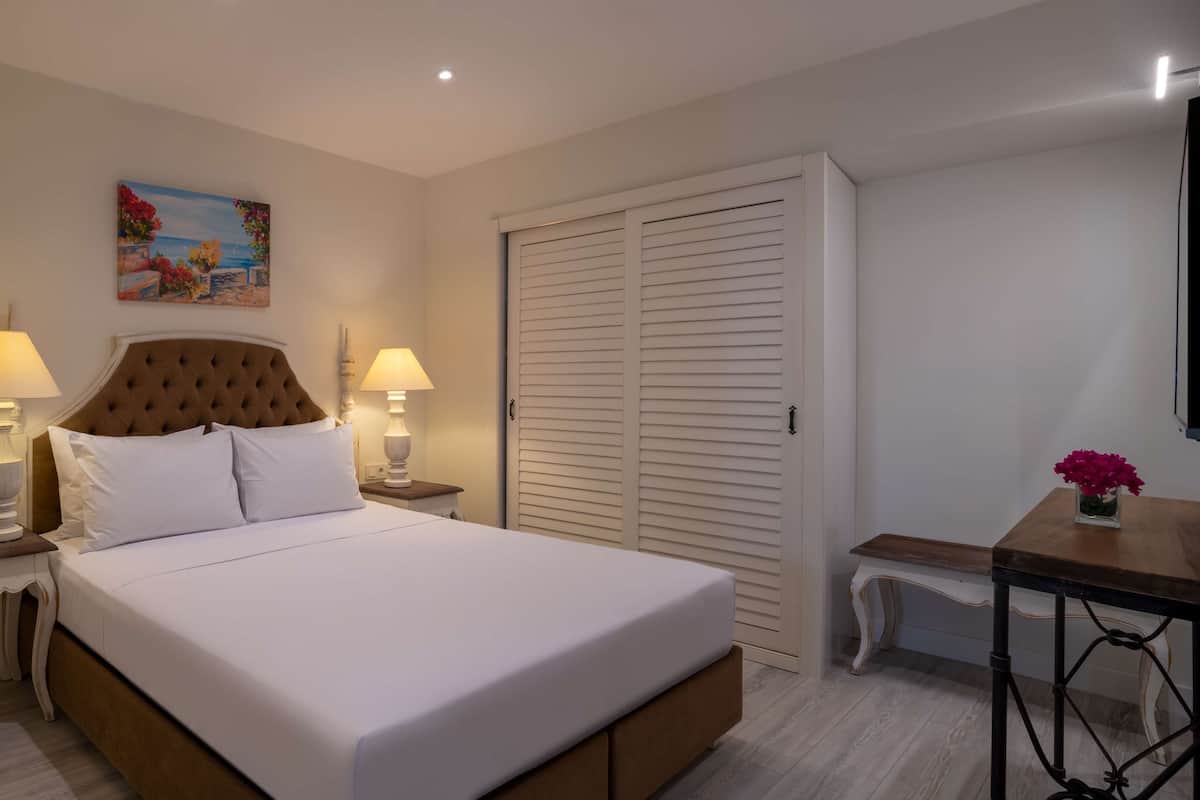 Un dormitorio ordenado con una cama grande, dos lámparas de noche, un cuadro en la pared, un armario cerrado y una mesita con un jarrón de flores.