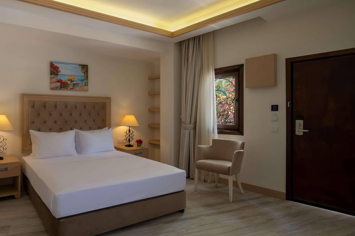 Una habitación de hotel con una cama grande, dos mesitas de noche con lámparas, una silla, una ventana con cortinas y una puerta.