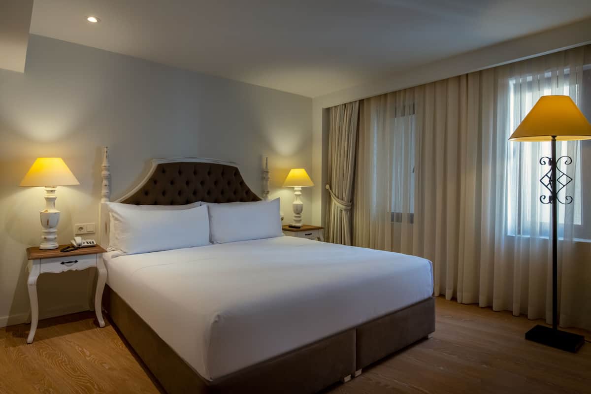 Habitación de hotel con una cama grande, dos mesitas de noche con lámparas, una lámpara de pie y un ventanal con cortinas.
