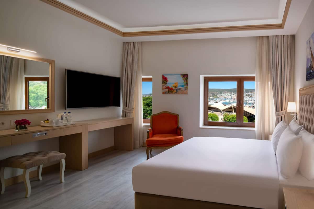 Habitación de hotel con una cama grande, un escritorio con televisor de pantalla plana, una silla y ventana con vista al puerto deportivo.