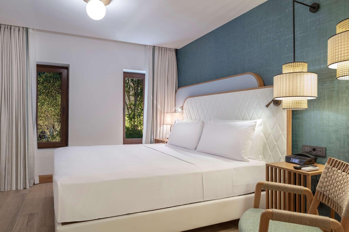 A modern hotel room with a large bed, a flat-screen TV, a desk with a chair, and a view of greenery through the windows.