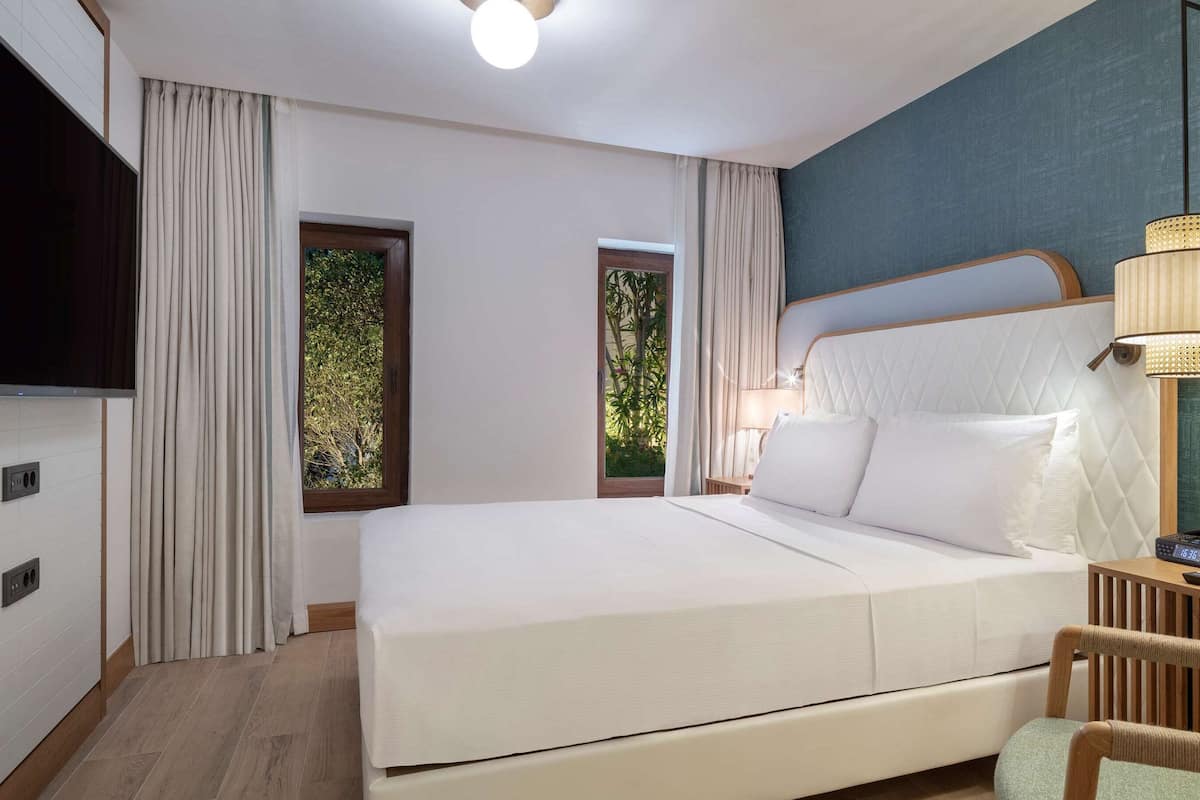 Habitación de hotel moderna con una cama grande, televisión de pantalla plana, escritorio con silla y vistas al verdor por las ventanas.