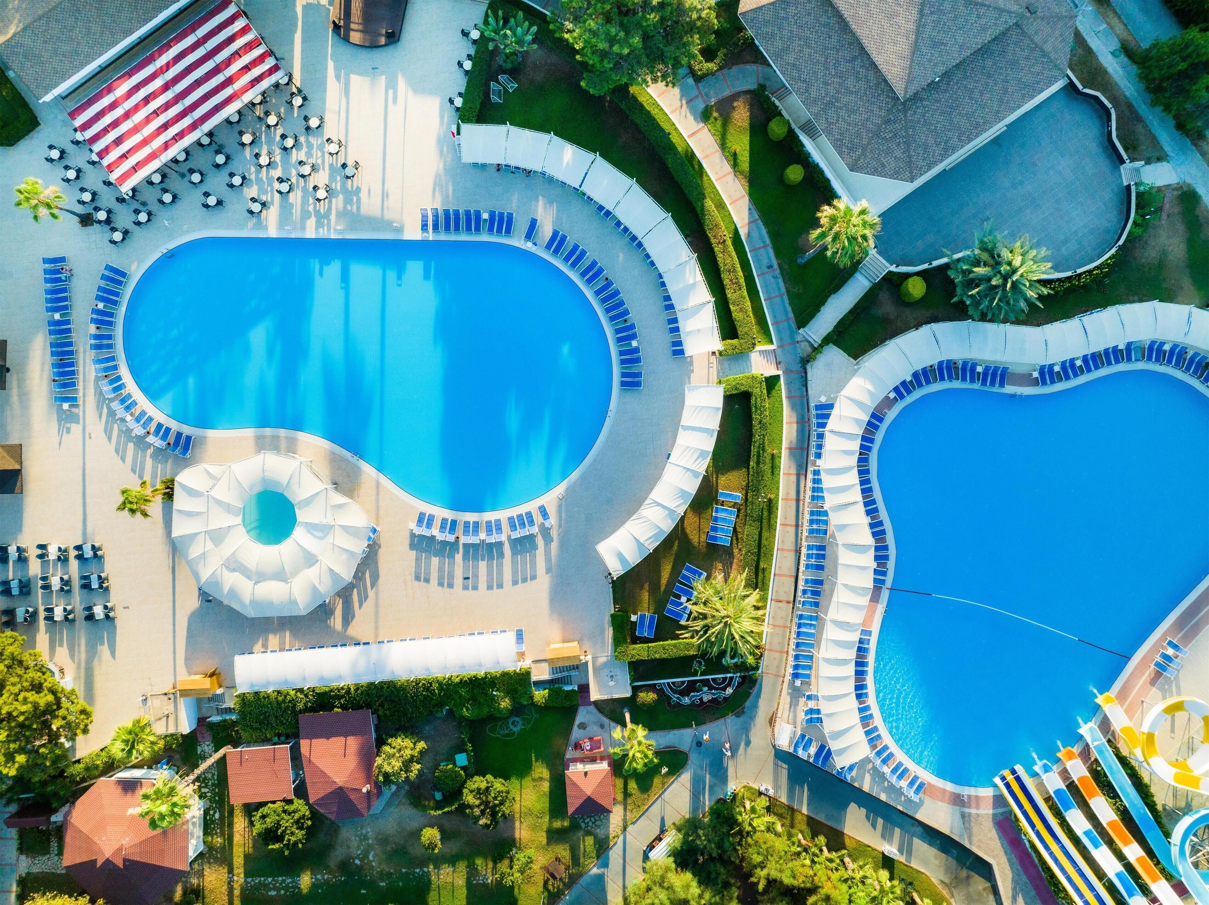 An aerial view of a resort with two large swimming pools, sun loungers, umbrellas, and a water slide.