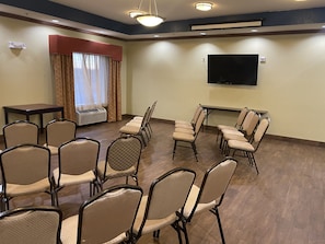 Uma sala de conferências com fileiras de cadeiras, uma mesa, um aparelho de ar condicionado, uma televisão na parede e uma janela com cortinas.