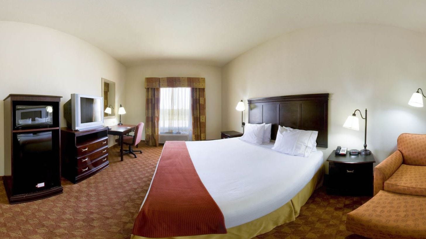 Quarto de hotel com uma cama grande, uma escrivaninha com televisão, uma cadeira, uma mesa de cabeceira com abajur e uma janela com cortinas.