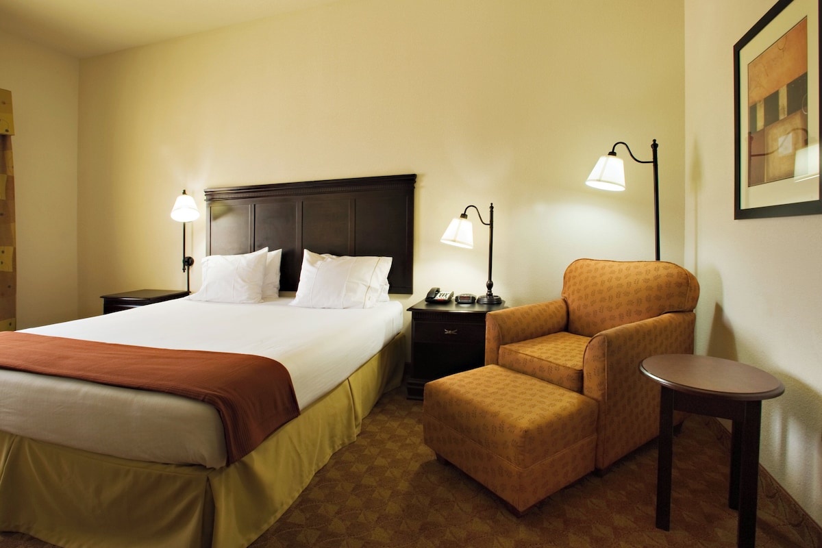 Quarto de hotel com uma cama grande, duas mesas de cabeceira, uma cadeira, uma mesinha e duas luminárias de chão.