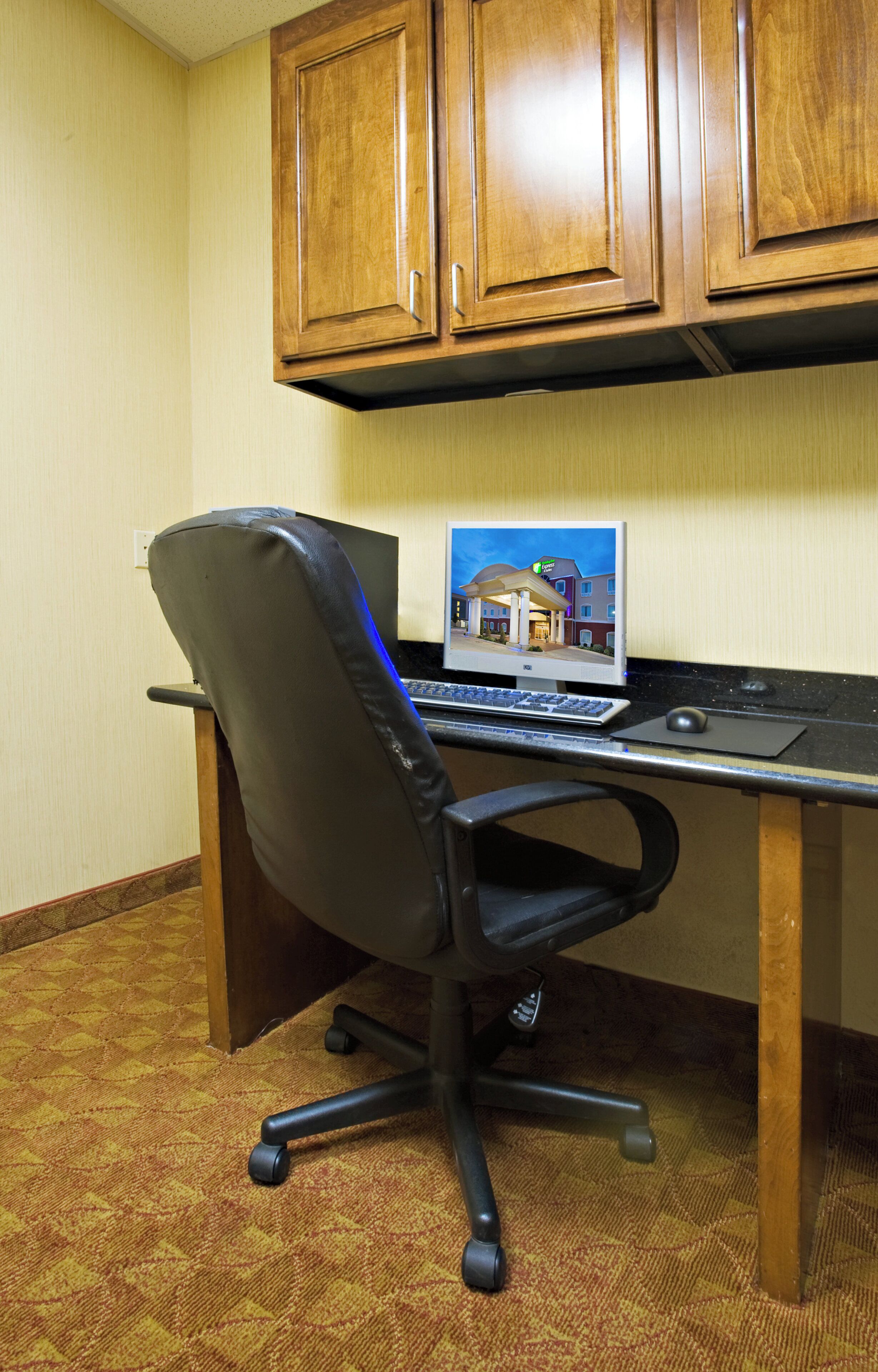A hotel room with a wooden desk, a black office chair, and a laptop displaying a hotel image.