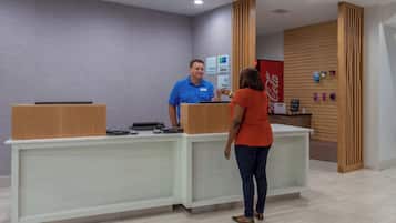 Um balcão de recepção com um homem e uma mulher, um letreiro da Coca-Cola e uma parede com vários avisos.