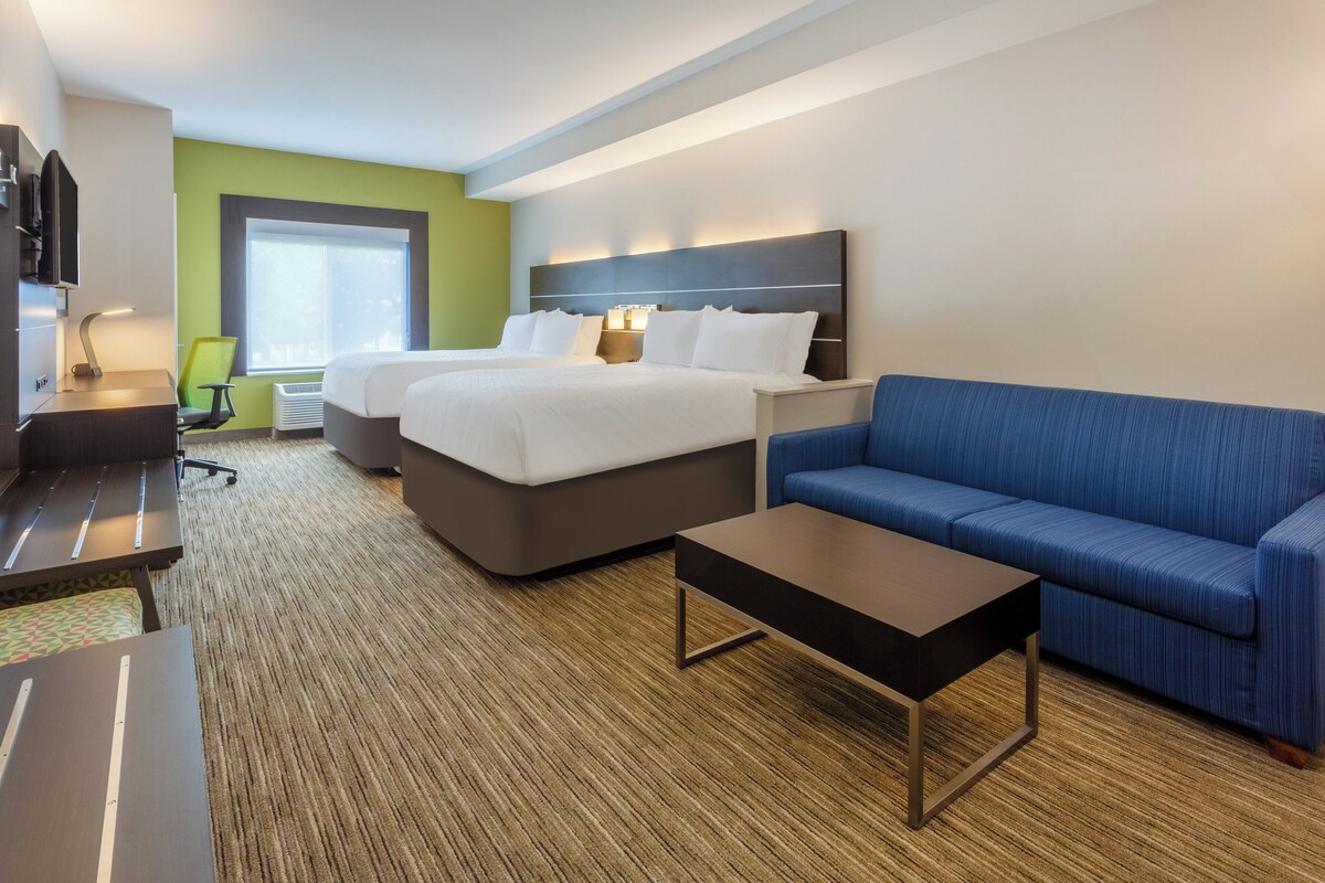 Quarto de hotel com duas camas, um sofá azul, uma mesa de centro, uma escrivaninha com cadeira e uma televisão.