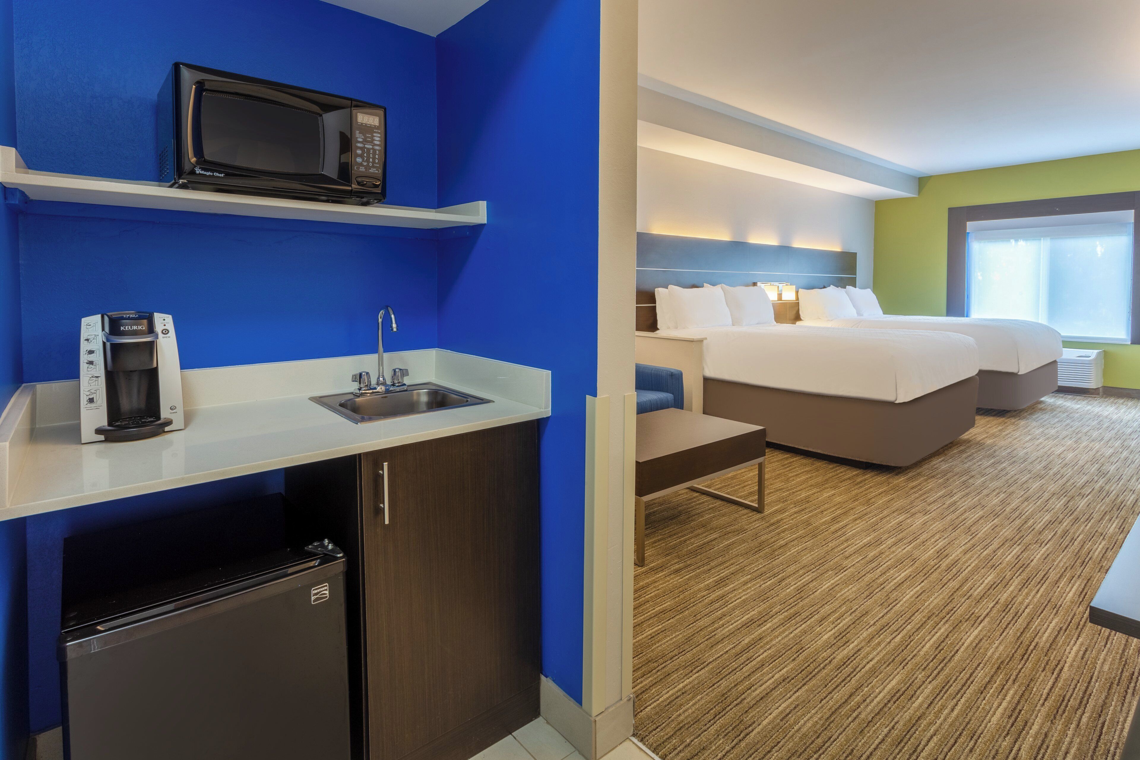 A hotel room with a kitchenette, a bed, a desk, and a window.