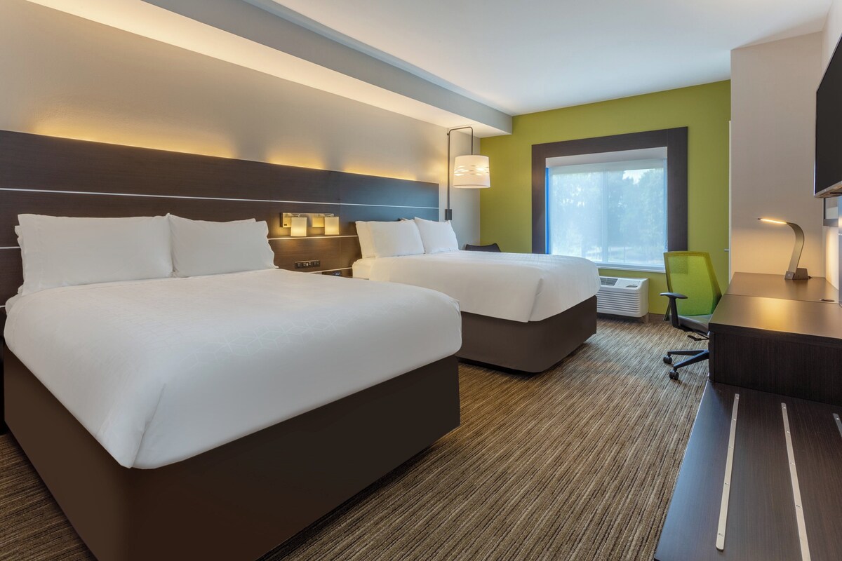 Quarto de hotel com duas camas, uma escrivaninha, uma cadeira, um abajur e uma janela.