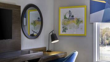 A modern room with a desk, a blue chair, a round mirror, a framed artwork, and a window with a view.